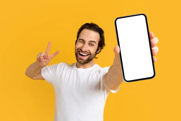 Cheerful emotional young european guy in white t-shirt show phone with empty screen and peace sign — Photo