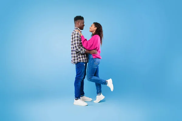 Tampilan samping pasangan muda Afrika Amerika yang bahagia dengan pakaian kasual berpelukan dan saling memandang dengan latar belakang biru — Stok Foto