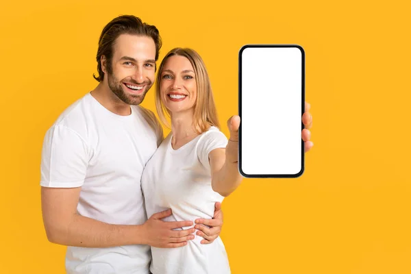 Smiling millennial european guy and lady in white t-shirts hugging, show smartphone with empty screen — Photo