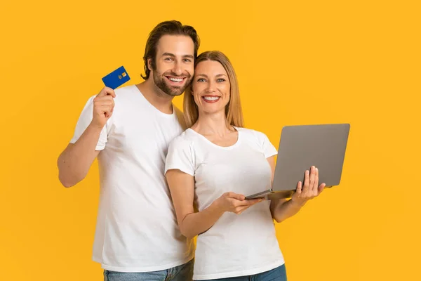 Smiling young caucasian man and woman in white t-shirts showing credit card and laptop — Stock Fotó