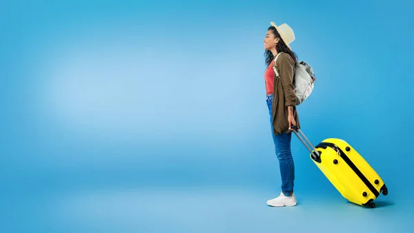 Zijaanzicht van millennial zwarte vrouw met rugzak en gele koffer op zoek naar lege ruimte op blauwe studio achtergrond — Stockfoto