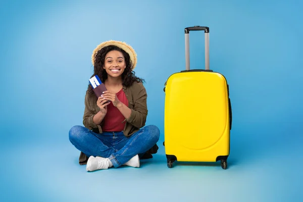 Young black woman sitting next to bright suitcase, holding international passport and plane tickets, smiling at camera — Stock fotografie