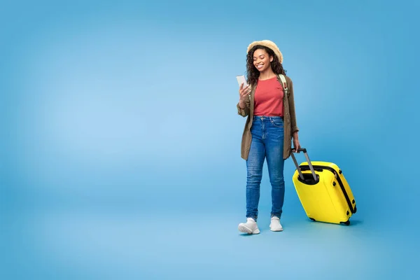 Young black woman with smartphone booking hotel or plane tickets online, walking with suitcase on blue studio background