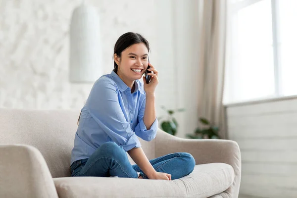 Positive korean lady sitting on sofa, talking on phone — Stockfoto