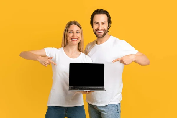 Χαμογελώντας χιλιετή Ευρωπαϊκό ζευγάρι σε λευκό t-shirts δείχνουν τα δάχτυλα στο laptop με λευκή οθόνη — Φωτογραφία Αρχείου