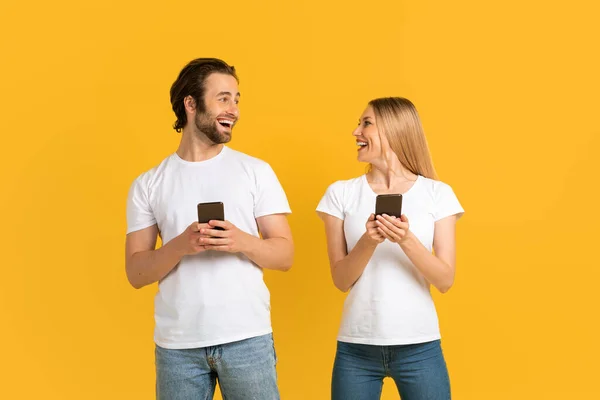Fröhliche Millennial-Kaukasier in weißen T-Shirts schauen einander an, halten Telefone in der Hand. — Stockfoto