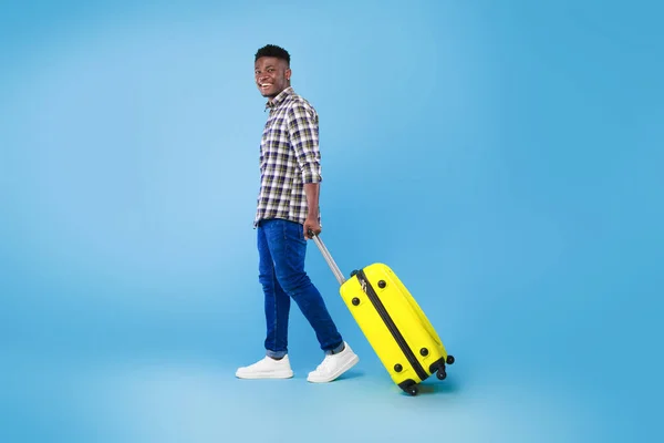 Happy young African American man walking with bright suitcase, going on summer vacation over blue background — Stockfoto