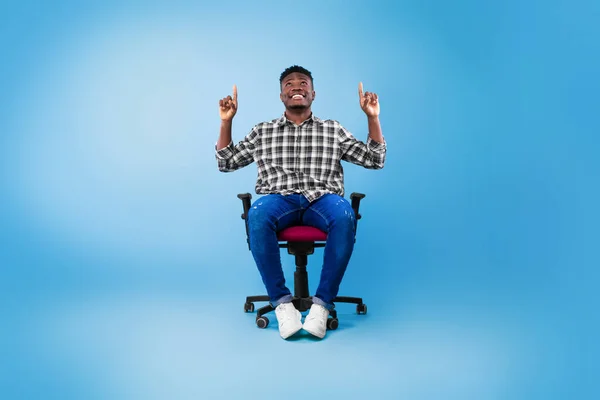 Atractivo joven negro apuntando hacia arriba en el espacio de copia, sentado en la silla sobre el fondo azul, maqueta para publicidad — Foto de Stock