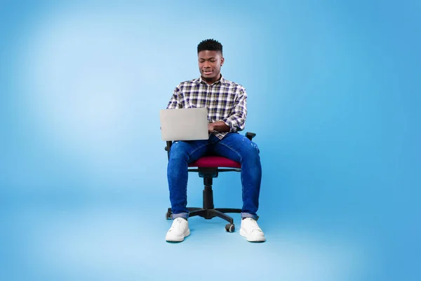 Dizüstü bilgisayarı olan genç siyahi bir adam sandalyede oturuyor, çalışıyor ya da internetten mavi stüdyo geçmişi üzerinde çalışıyor. — Stok fotoğraf