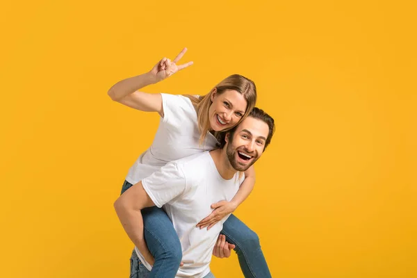 Smiling millennial european man and woman in white t-shirt take wife on back and have fun, lady shows peace sign — Stok fotoğraf