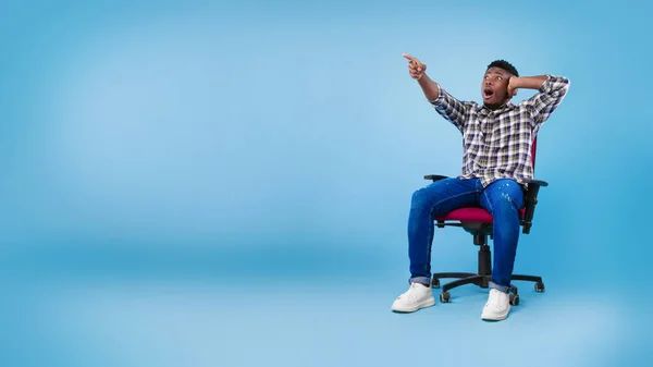 Shocked young black man sitting on chair, pointing at empty space for your advertisement on blue studio background — Stok fotoğraf