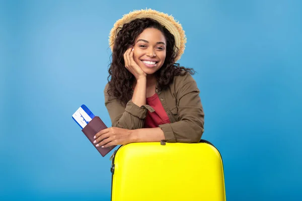 Mooie jonge zwarte dame met paspoort en vliegtickets, leunend op felgele koffer over blauwe achtergrond — Stockfoto