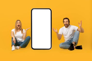 Neşeli Avrupalı genç adam ve ağzı açık kadın ellerini kaldırıp kocaman bir telefonla yerde oturuyorlar.