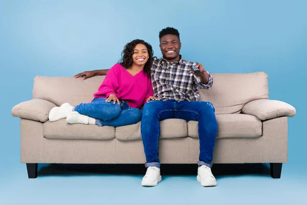 Happy young black couple with remote control sitting on sofa and watching TV over blue studio background, full length — Stockfoto