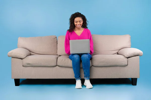 Glad ung svart kvinna i casual wear arbetar online, sitter på soffan och använder laptop på blå studio bakgrund — Stockfoto