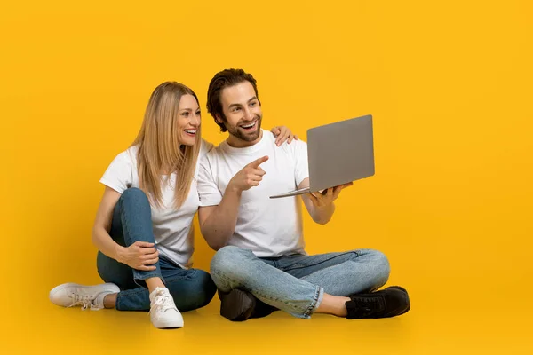 Χαμογελώντας νεαρός Ευρωπαίος με γενειάδα και κυρία κάθονται στο πάτωμα, δείχνουν δάχτυλο στο φορητό υπολογιστή απομονώνονται σε κίτρινο φόντο — Φωτογραφία Αρχείου