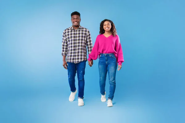 Penuh panjang milenial Afrika Amerika pasangan berjalan, berpegangan tangan, tersenyum pada kamera di latar belakang studio biru — Stok Foto