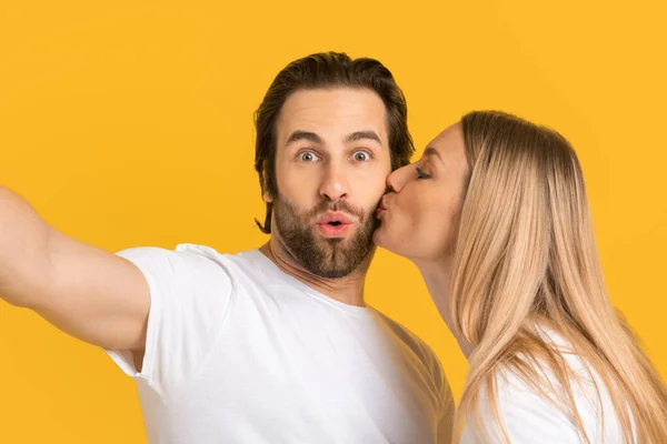 Jovem mulher loira europeia feliz em branco t-shirt beijos marido surpreso na bochecha, isolado no fundo amarelo — Fotografia de Stock