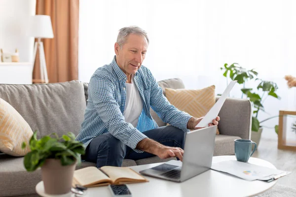 Άντρας κρατώντας χαρτί ανάγνωση έκθεση εργασίας σε φορητό υπολογιστή στο σπίτι — Φωτογραφία Αρχείου