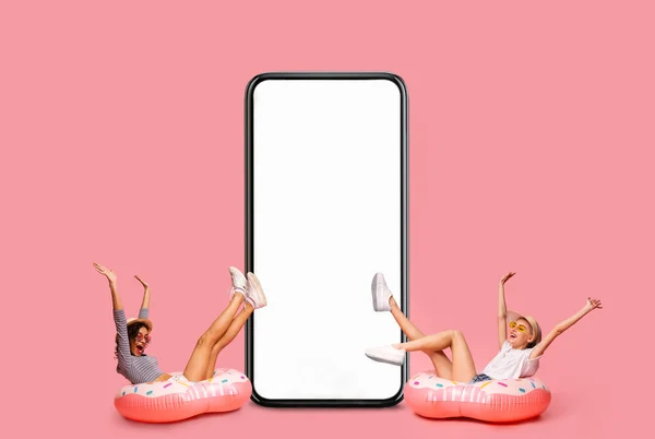Cheerful Young Women Sitting In Inflatable Pool Rings Next To Blank Smartphone — Stockfoto