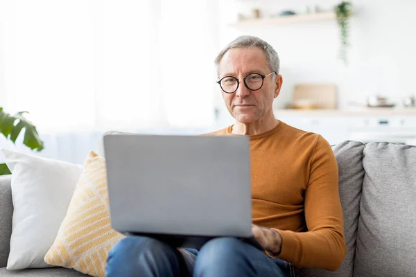 Magabiztos alkalmi érett férfi szemüvegben laptoppal otthon — Stock Fotó