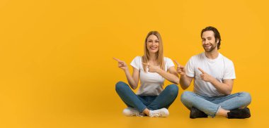 Smiling young european guy with beard and woman sit on floor, point fingers at empty space