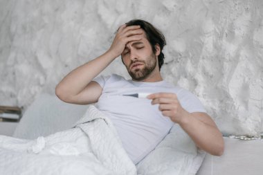 Despaired millennial caucasian guy with stubble on bed, touches forehead, looks at thermometer in bedroom