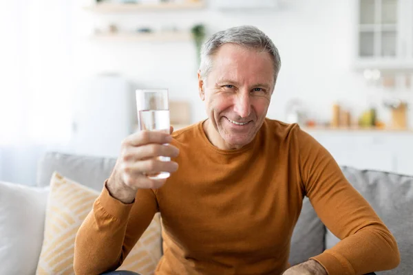 Smiling mature man sitting on couch holding glass of water — Stock Photo, Image