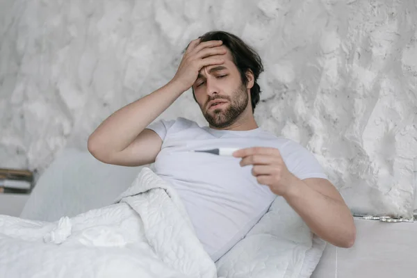 Απελπισμένος άντρας της χιλιετίας με γένια στο κρεβάτι, αγγίζει το μέτωπο, κοιτάζει το θερμόμετρο στην κρεβατοκάμαρα. — Φωτογραφία Αρχείου