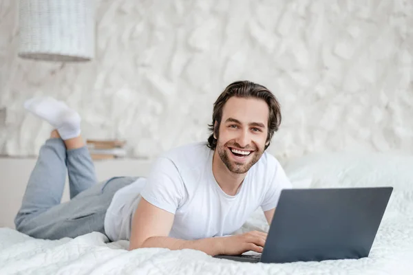 Glad young caucasian man with stubble lies on bed with computer, surfing in internet in light bedroom interior — Foto de Stock