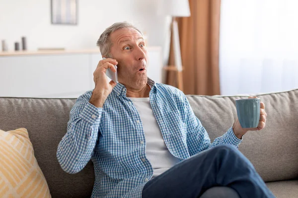 Shocked mature man talking on cellphone sitting on sofa — стоковое фото