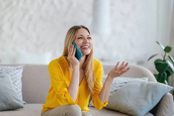 Sonriente joven rubia europea con ropa amarilla se sienta en el sofá, llama por teléfono y gesticulando con su mano — Foto de Stock