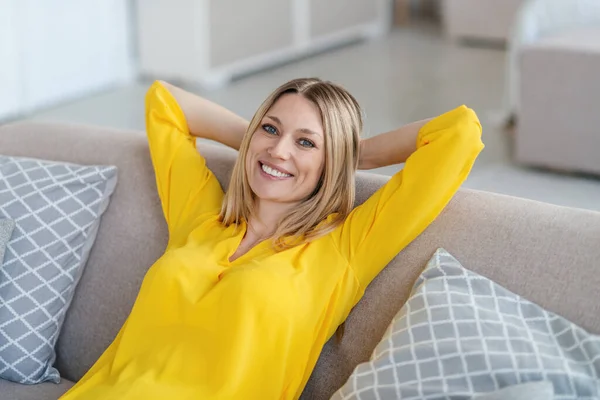 Cheerful pretty young blond european woman in yellow clothes sits on sofa with hands behind her head — 图库照片