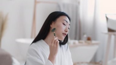 Young attractive asian woman making self massage with natural stone massager, sitting at bathroom at home