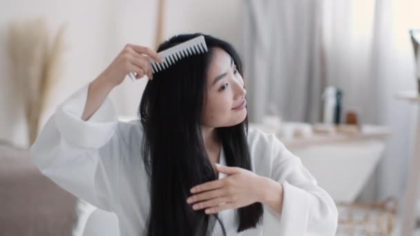 Bellezza di routine. Giovane signora asiatica spensierata che indossa accappatoio spazzolando i suoi lunghi capelli setosi, seduto in bagno, spazio libero — Video Stock