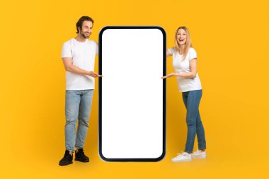 Beyaz tişörtlü mutlu beyaz erkek ve kadın boş ekranlı büyük bir telefon sunar.