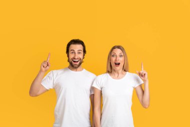 Glad excited millennial european couple in white t-shirts with open mouths show fingers up to empty space or got idea