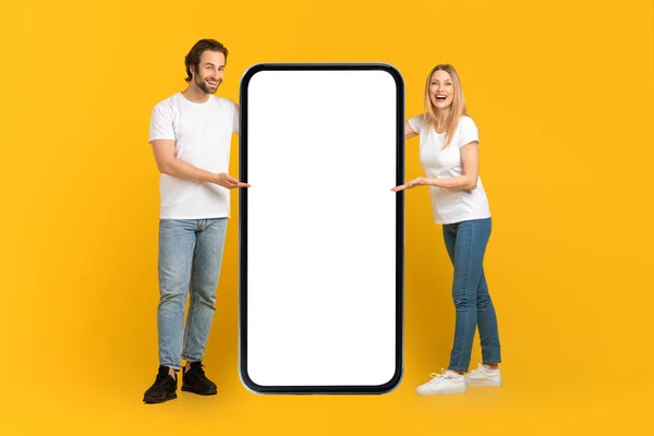 Happy young caucasian male and female in white t-shirts presentation huge phone with empty screen — Stok fotoğraf