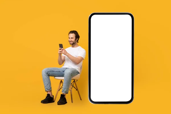 Sorrindo jovem europeu macho em branco t-shirt senta-se na cadeira, digitando no telefone perto enorme smartphone — Fotografia de Stock