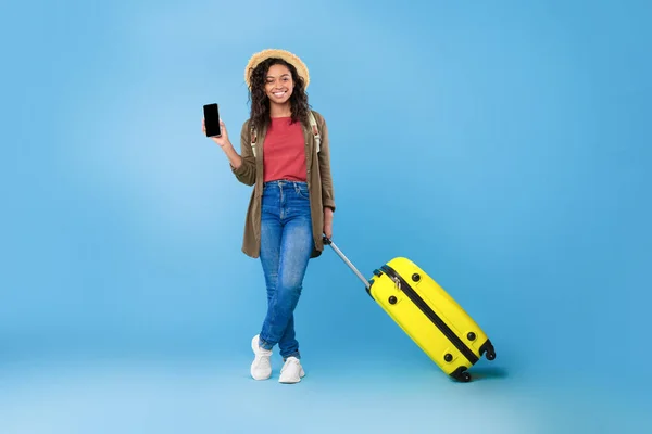 Young black woman with suitcase showing smartphone with blank screen, offering mockup, booking summer vacation online — Stockfoto