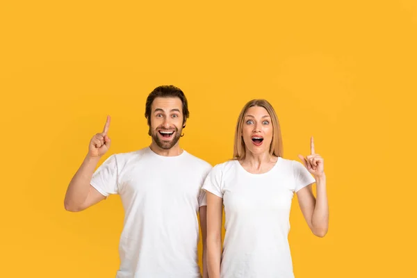 Feliz casal europeu milenar animado em camisetas brancas com bocas abertas mostrar dedos até espaço vazio ou tem ideia — Fotografia de Stock