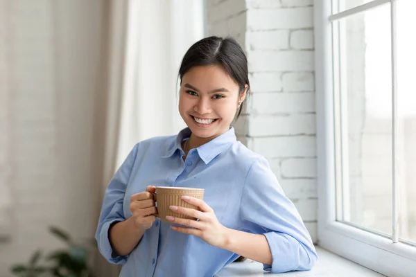 Positive pretty asian woman standing by window, drinking coffee — Foto de Stock