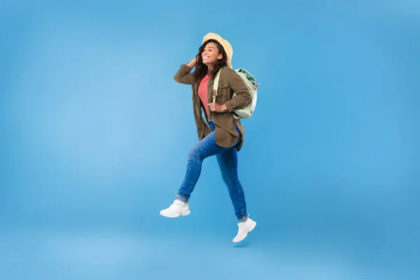 Plná délka mladých Afroameričanky cestovatel běží s batohem, skákání ve vzduchu přes modré pozadí — Stock fotografie