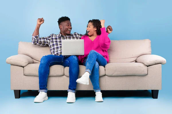 Opgewonden millennial zwart paar zitten op de bank met laptop, het verhogen van de handen omhoog, vieren van succes of enorme overwinning — Stockfoto