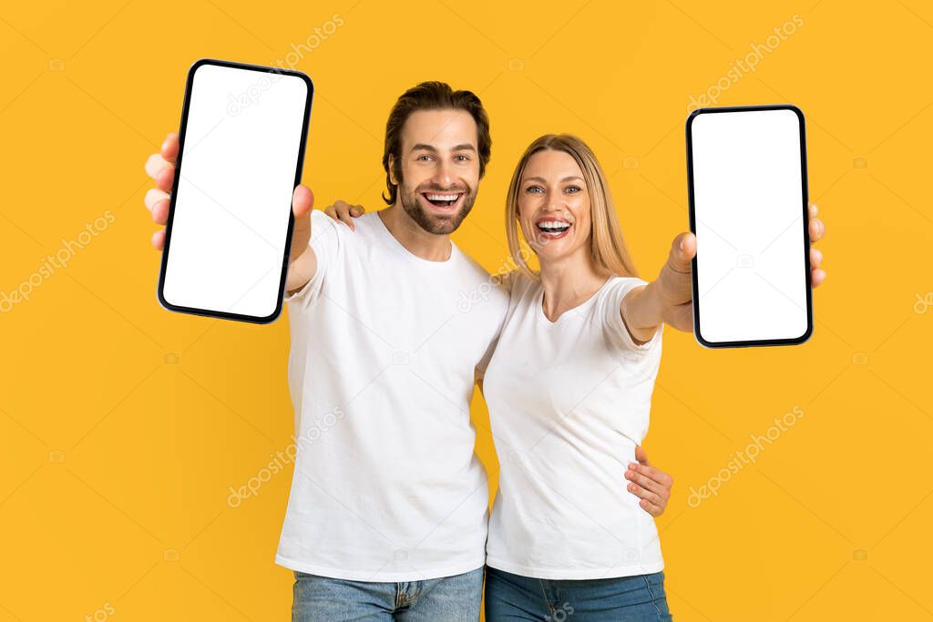 Cheerful young european wife and husband in white t-shirts show phones with blank screen