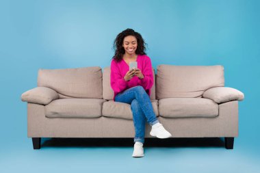 Happy millennial black lady using cellphone, working or studying remotely, sitting on sofa over blue studio background