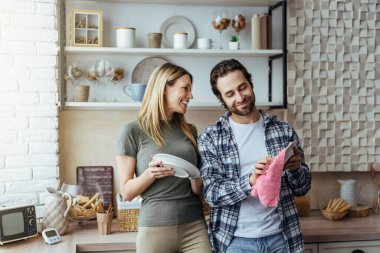 Happy european millennial man with stubble and blond wife wash dishes, wipe plates in light kitchen interior
