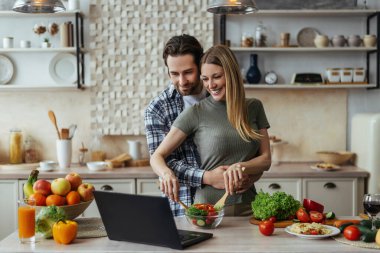 Gülümseyen koca sakallı, Avrupalı milenyum eşi, kadın salata hazırlıyor ve dizüstü bilgisayara bakıyor.