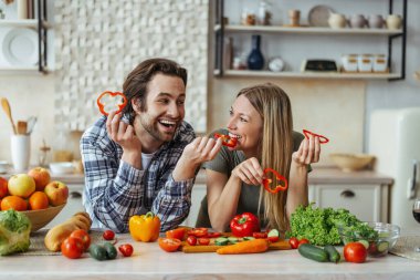 Kirli sakallı mutlu Avrupalı genç adam biber sarışını besliyor, eğleniyor, organik sebzelerle masada.