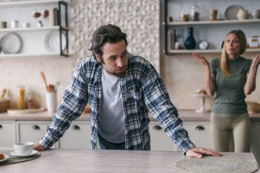 Sad dissatisfied european millennial male with stubble sighs, upset blonde wife screams hasband